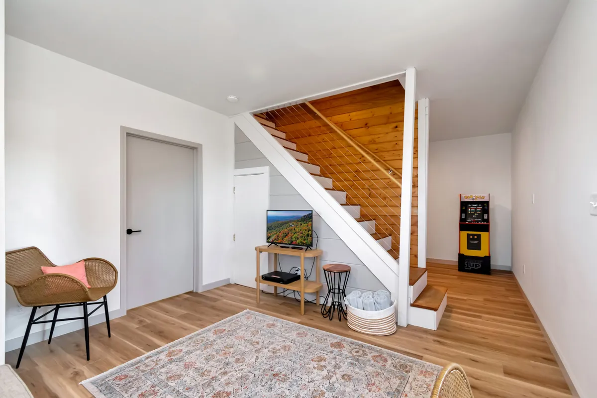 The interior scene includes a staircase, a TV stand with a large screen, an area rug, an arc chair, and a classic arcade machine. The setting blends cozy seating for movie nights with gaming, all within an open, multi-use family space.