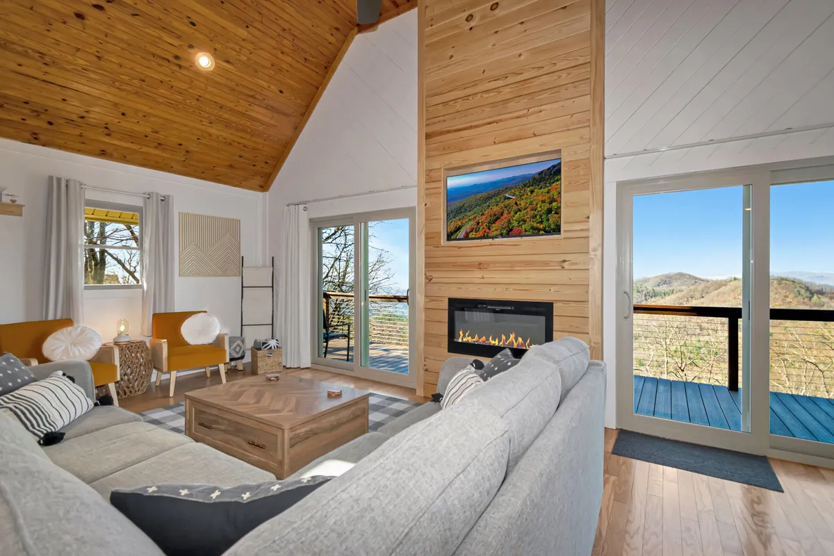 A welcoming living space featuring a stone fireplace, large windows that frame the scenic outdoor view, a wooden accent wall, and comfortable seating arranged for conversation or relaxation. The room blends rustic charm with modern touches, creating a cozy yet stylish environment.