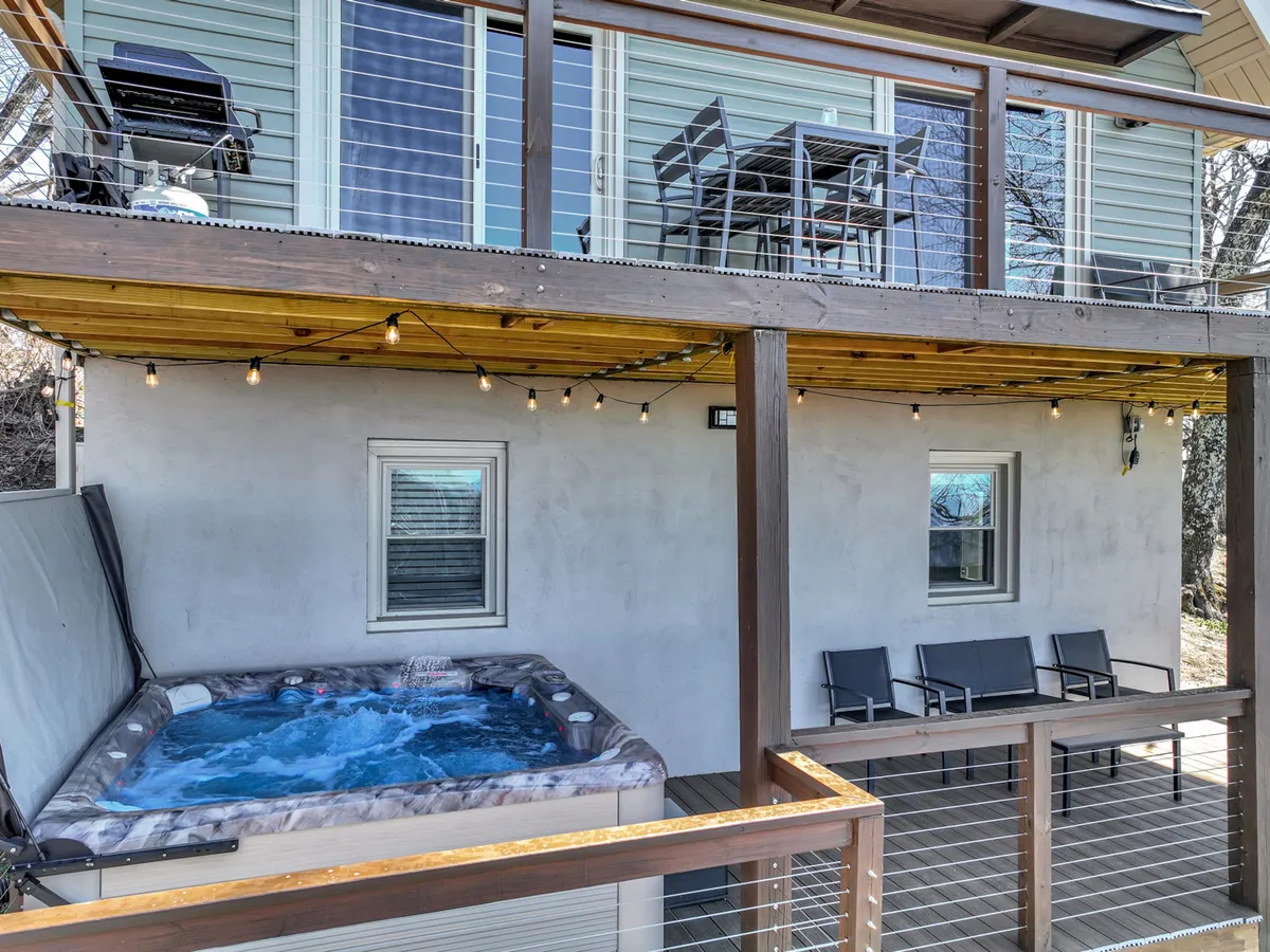 This image captures a modern hot tub on a spacious deck, complemented by a cozy outdoor seating area and a balcony dining set. The backdrop features tranquil mountain scenery and inviting string lights, evoking relaxation and a sense of retreat amidst nature.