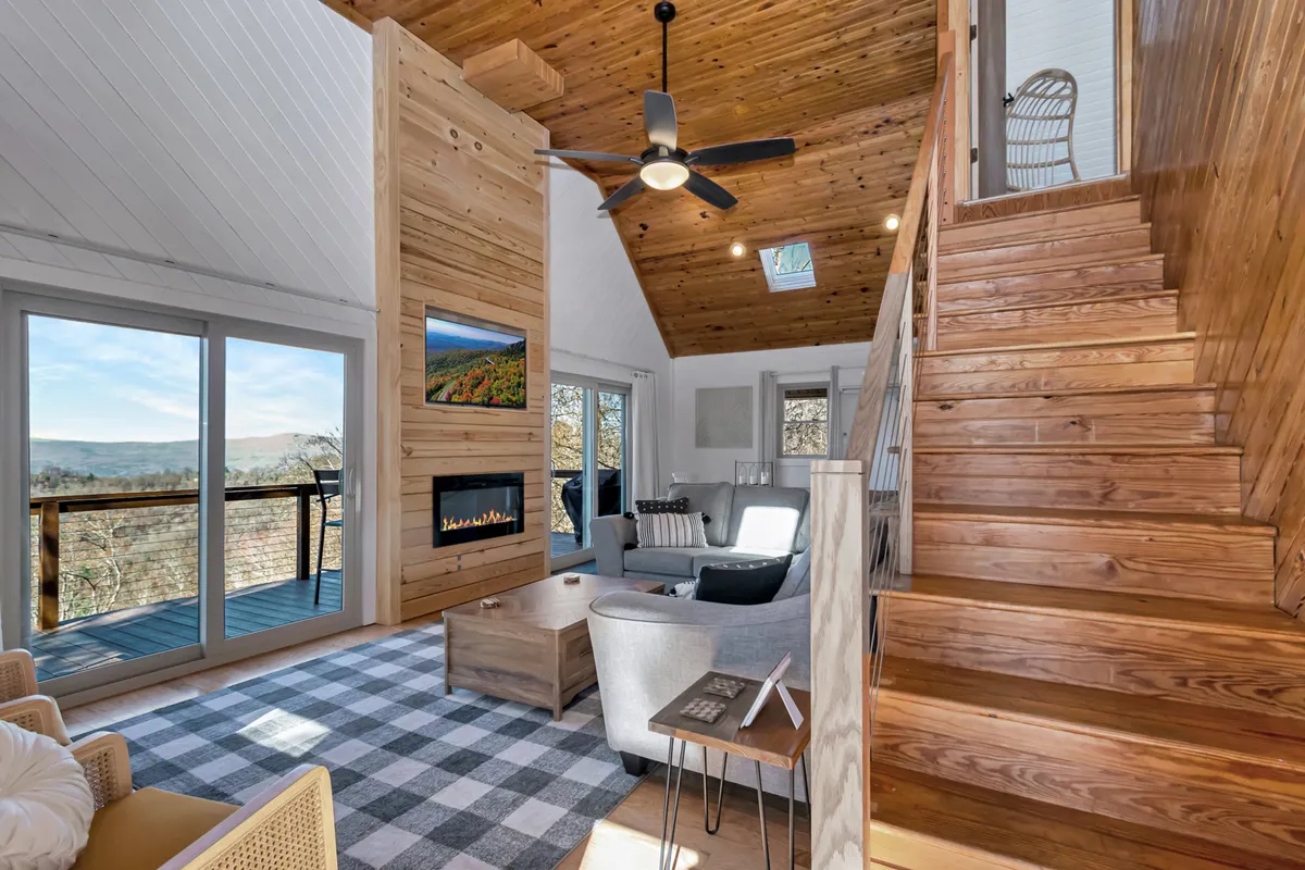 A spacious living area with a wooden accent wall, large windows offering beautiful mountain views, a modern fireplace, and a comfortable seating arrangement. The area is flooded with natural light and features a staircase leading to upper levels, creating an open and inviting atmosphere.