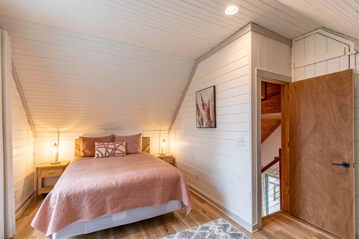 A cozy bedroom with a plush Queen bed dressed in a pink bedspread, complemented by nightstands with glowing lamps and art on the wall. An open door reveals a staircase, adding to the retreat-like feel.