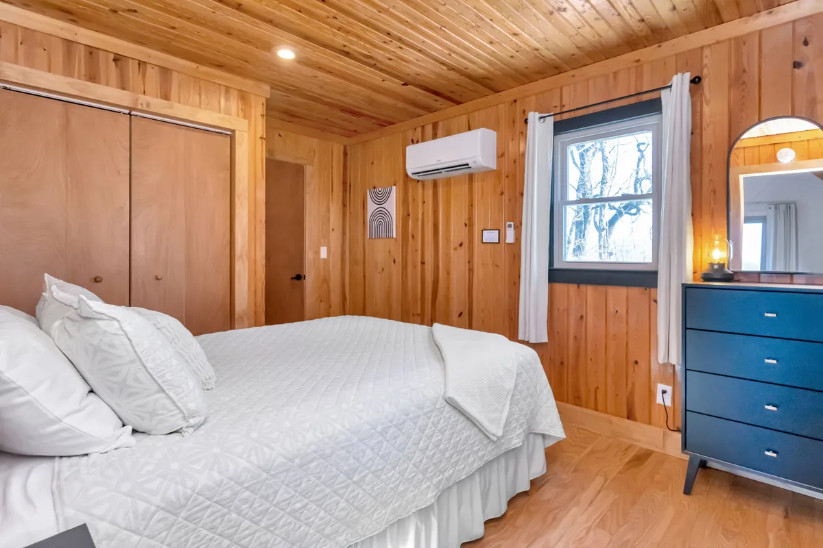 A tranquil bedroom featuring a quilted Queen bed, dark dresser, window with gentle curtains, and calming wall art. Sunlight softly fills the space, evoking relaxation and coziness.