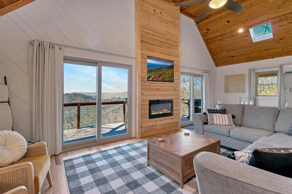 The image showcases a cozy living area with floor-to-ceiling windows framing breathtaking mountain views, a warm wooden feature wall with a crackling fireplace, plush seating, and a central coffee table—creating an inviting space perfect for relaxation or gathering with loved ones.